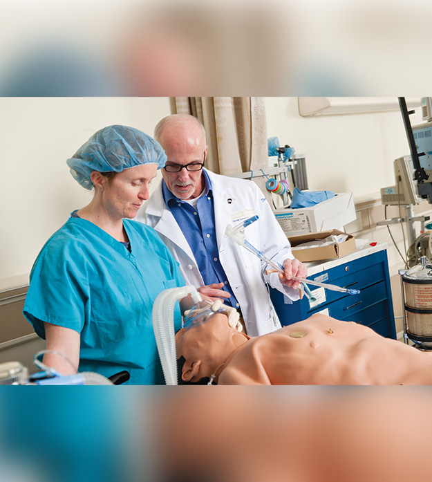  A nurse holds a mask over the mouth of a human test dummy to practice administering anesthesia while a physician monitors their steps.