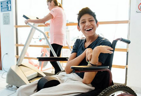 Photo of a smiling boy in a wheelchair and girl walking on a treadmill.