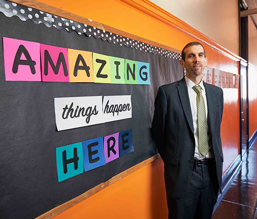 Andrew Lukov stands in a suit and tie in front of a sign that says “amazing things happen here” in the bright orange hallways of Southwark School.
