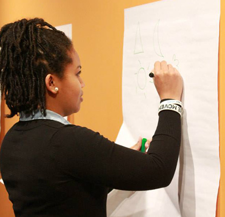 A student wearing a long-sleeved black shirt draws on a large white piece of paper hung from an orange wall.