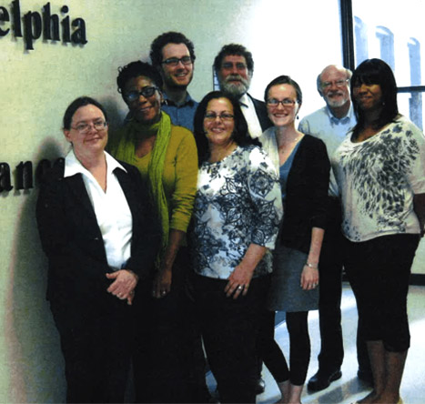 A group of PLA employees pose together wearing business casual clothing while smiling at the camera in front of a wall that reads “Philadelphia Legal Assistance.”