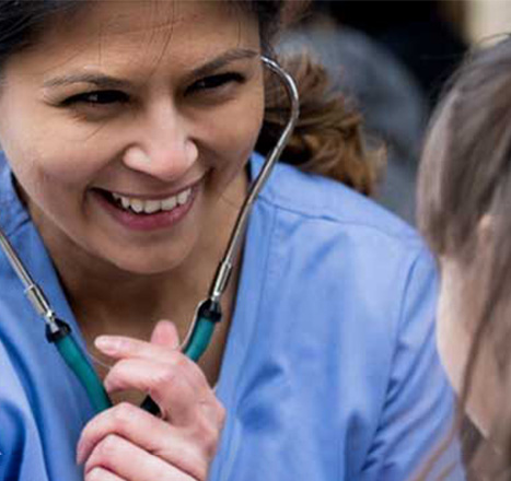 A woman wearing blue scrubs and a stethoscope smiles at a young girl as she provides patient care.