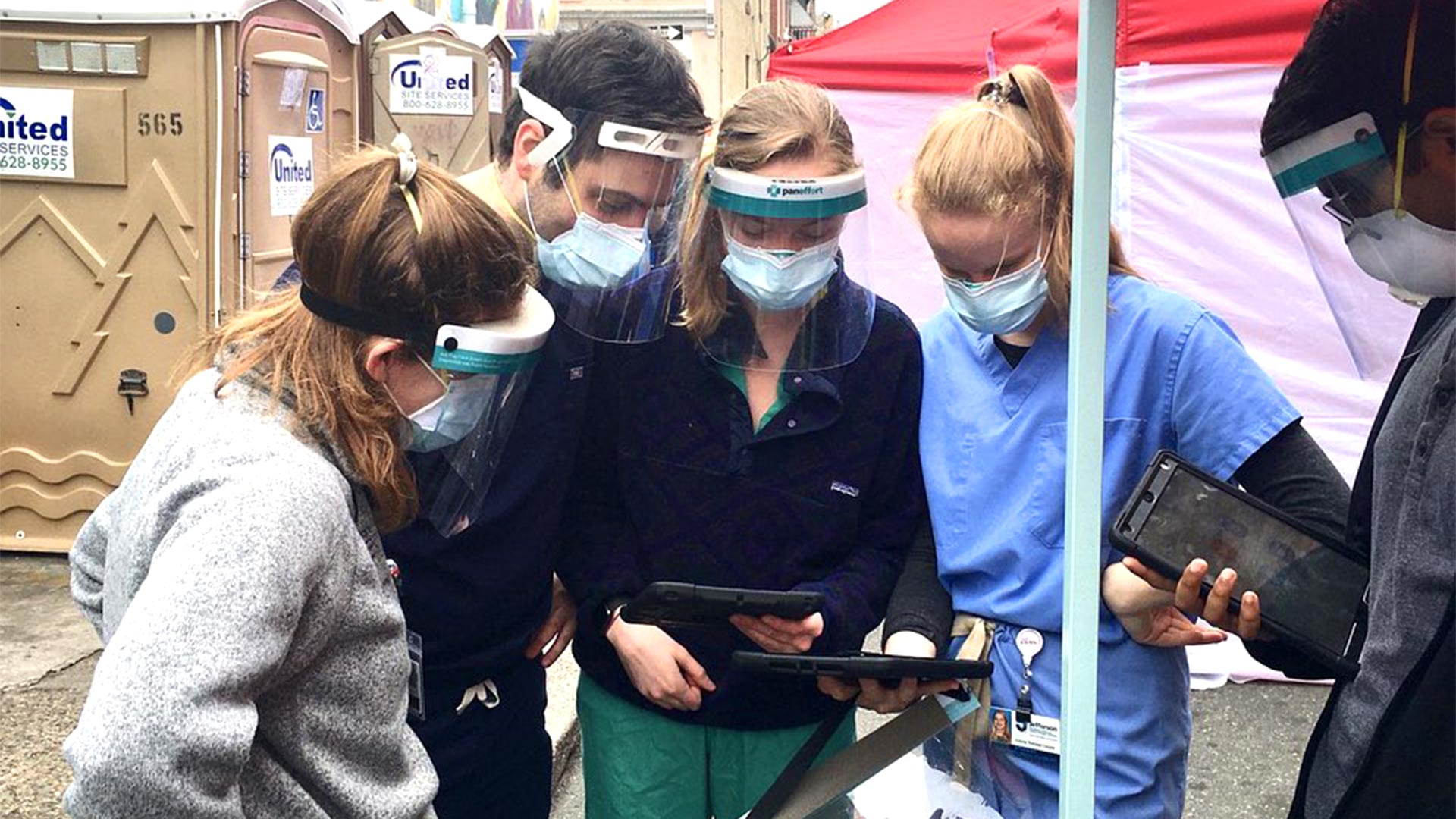 A group of JeffHOPE workers in scrubs, face masks and face shields stand at an outdoor clinic looking at tablets.