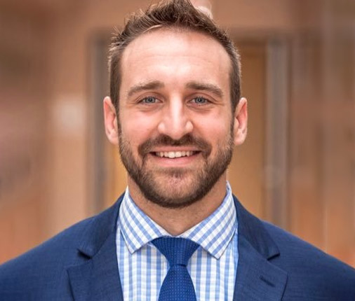Dr. Alex Hajduczok wears a navy suit with a collared shirt in a blue checkered pattern, a blue tie, and a smile as he poses for a professional headshot.