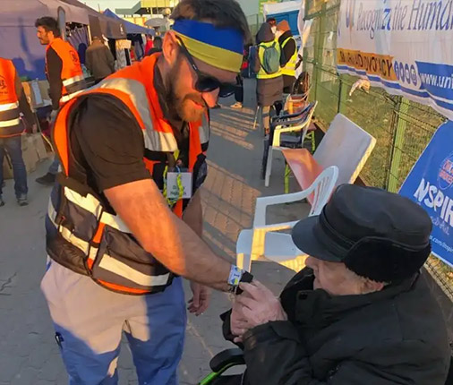 Dr. Alex Hajduczok stands with a headband the colors of the Ukrainian flag, a reflective vest and scrubs while a patient in a wheelchair grasps his hand.
