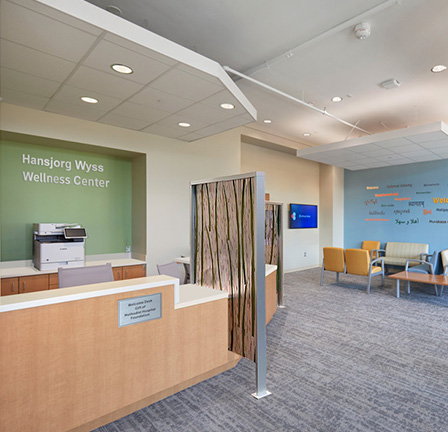 The interior of the Wyss Wellness Center lobby, which features bright blue and green walls that say the name of the center and welcome in several languages, a reception desk, and waiting room furniture including couches, chairs and shelves.