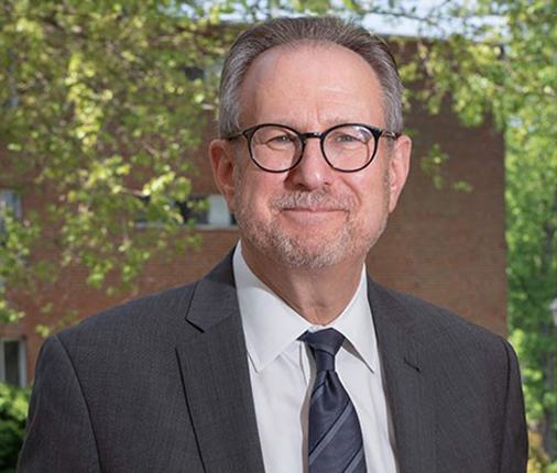 Dr. Matt Baker wears a suit, tie, and glasses while smiling at the camera in a professional headshot pose.