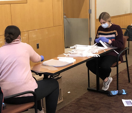 A group of students wearing face masks and gloves assemble face shields from various components stacked on top of tables spread around a room.