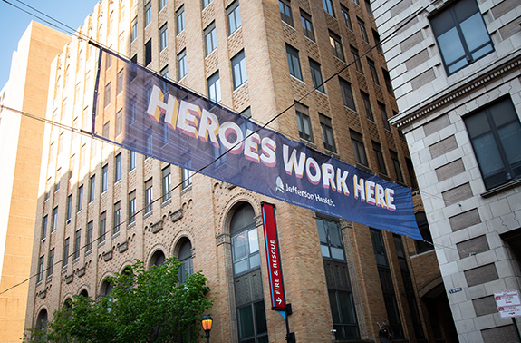 A banner hanging across two downtown buildings reads “Heroes work here” in all caps, white lettering on a branded blue Jefferson banner.