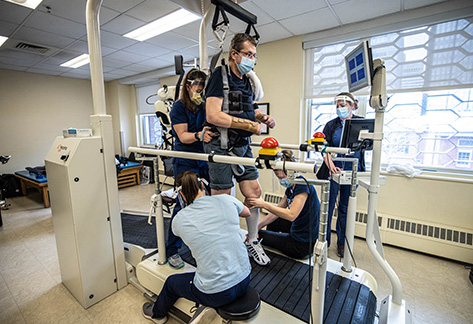 A person wearing a mask and secured into a device hanging above a treadmill is surrounded by health care workers wearing face masks and shields as they assist the patient. 