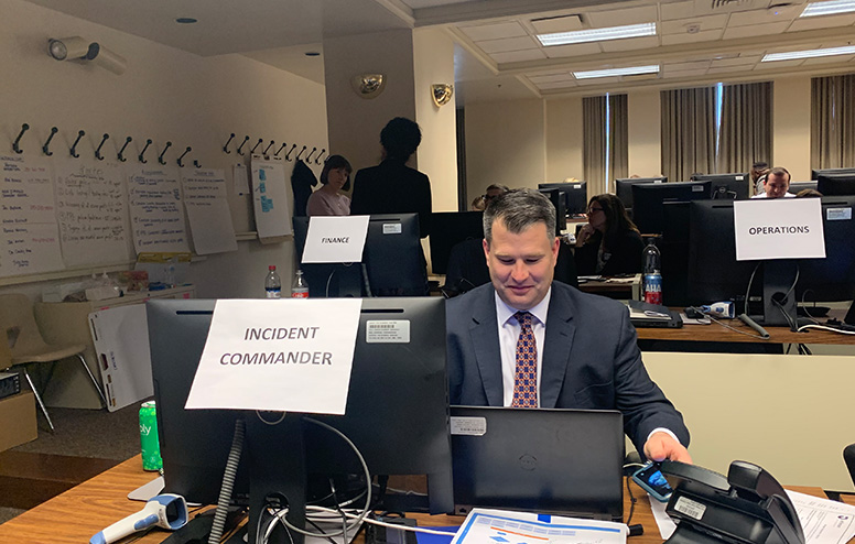 A group of people sit and stand around computers in a war room-type environment with signs that read finance, operations and incident commander.