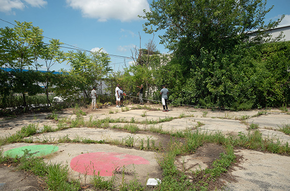 A group of Park in a Truck workers use a tape measure while making plans in an empty, overgrown city lot.