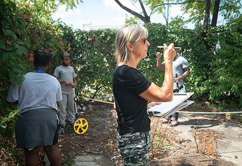 Kim Douglas holds a pen in one hand a clipboard in another while evaluating an empty city lot as other workers take measurements in the background.