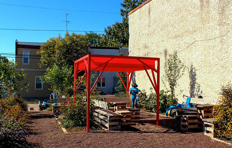 A completed red canopy provides shade over wooden benches surrounded by landscaping at a Park in a Truck installation.