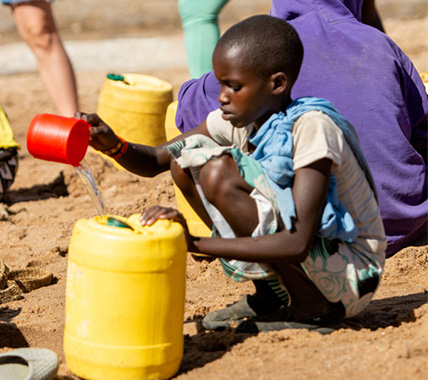 A child kneels while pouring water from a red cup into a large yellow container in front of a puddle of water on the ground.