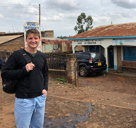 Michele Ganz wears a long-sleeved black shirt and jeans with a backpack slung over one shoulder while he stands in front of a building that reads, “Wema Hospital.”