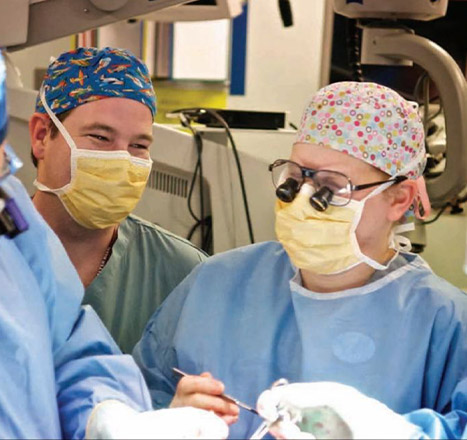 E.J. and Stephanie Caterson wear scrubs, surgical masks, and head coverings while in surgery surrounded by other hospital workers also in scrubs.