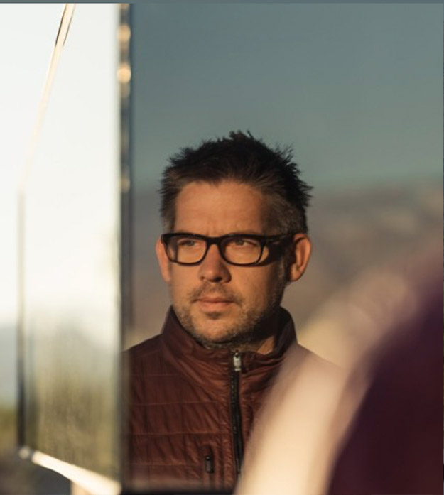 Sean Lockyer wears black-rimmed glasses and a maroon zip-up jacket while looking into the distance.