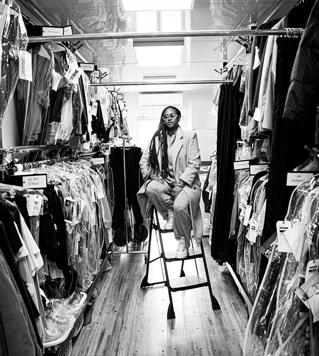 A black-and-white photo shows Charlese Antoinette sitting on a step ladder with clothing in clear plastic garment bags hanging on floor-to-ceiling racks on either side of her.