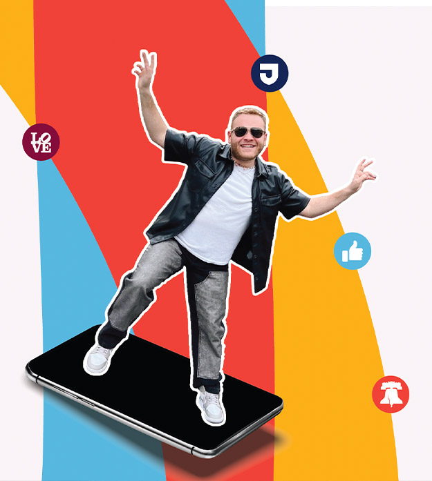 Brandon Edelman wears sunglasses, a short-sleeved black leather jacket, white T-shirt, jeans, and tennis shoes while making peace signs with both arms in the air as he’s depicted standing on a large smartphone against a colorful background with various logos/icons.