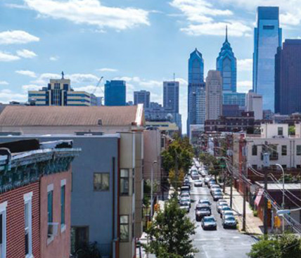 A view from North Philadelphia shows a long city block lined with cars, buildings and power lines as the Philadelphia skyline towers in the background. 