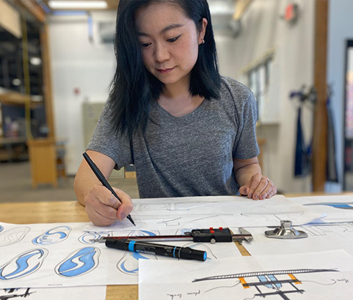Yuhan Zhang wears a short-sleeve, heather gray shirt while working on a Swell Surf Foot prosthetic sketch on a table covered with drawings.