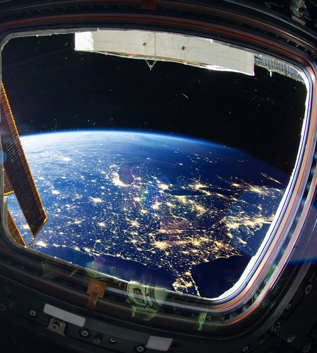 The view looking out two windows of a spacecraft shows Earth below, with lights visible throughout the populated areas of the United States.