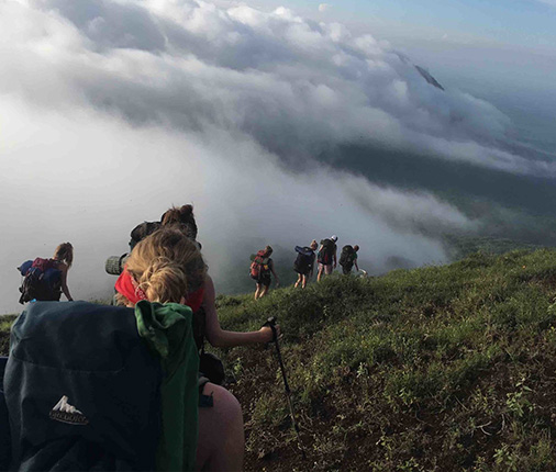 A group of students hiking on a grassy mountain above the clouds.