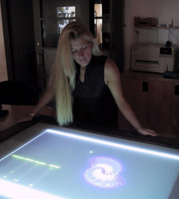 Dr. Lizbeth Goodman stands over a large digital display that lights up a dark room and depicts some kind of geographic spiral on the screen.