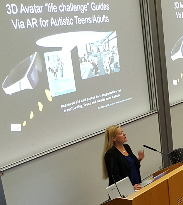 Dr. Lizbeth Goodman stands at a podium speaking into a microphone while a large projector screen behind her displays images and text about 3D avatar guides.