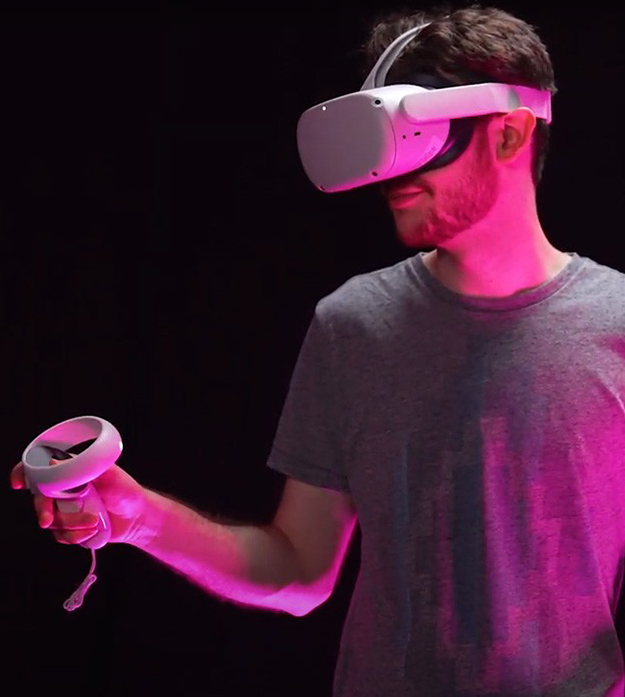 A person stands cast in pink light against a white background wearing a gray T-shirt, beard, and white virtual reality headset while holding a matching white remote in their right hand.