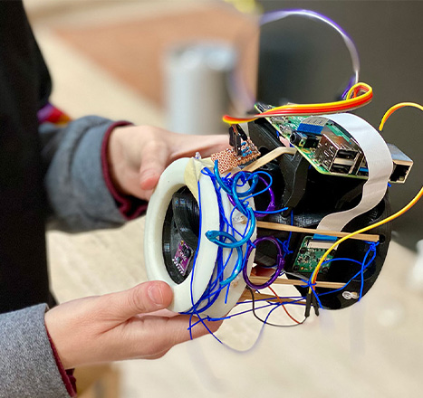 A pair of hands holds up an electronic device with exposed batteries and wiring of various colors.