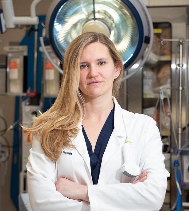 Dr. Patricia Henwood wears a white lab coat over a navy shirt as she stands in a laboratory with her arms crossed while looking at the camera.