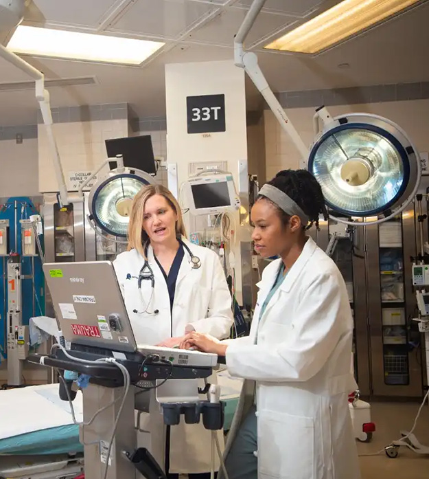 Dr. Patricia Henwood wears a white lab coat and stethoscope while looking at a computer in an operating room with a colleague who is also wearing a white lab coat.