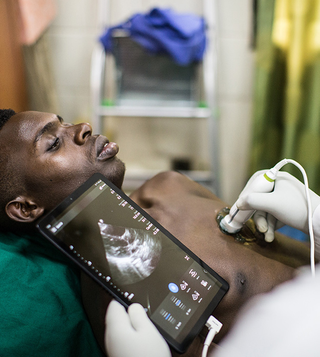 A doctor with their back turned to the camera uses the Philips Lumify with Reacts on a patient lying face-up in a bed at the University Hospital of Kigali, Rwanda.