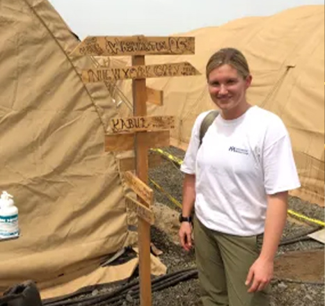 Dr. Patricia Henwood wears a white T-shirt and cargo pants while standing at a wooden crossroads sign while standing outside of large, beige canvas tents.