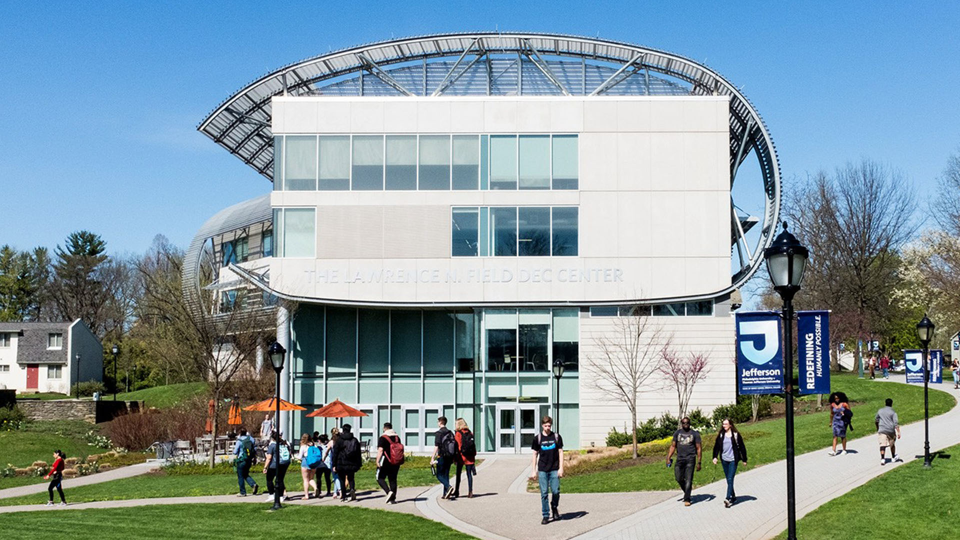 Several students walk on the sidewalk through a grassy lawn in front of a gray building labeled the Lawrence N. Field DEC Center.