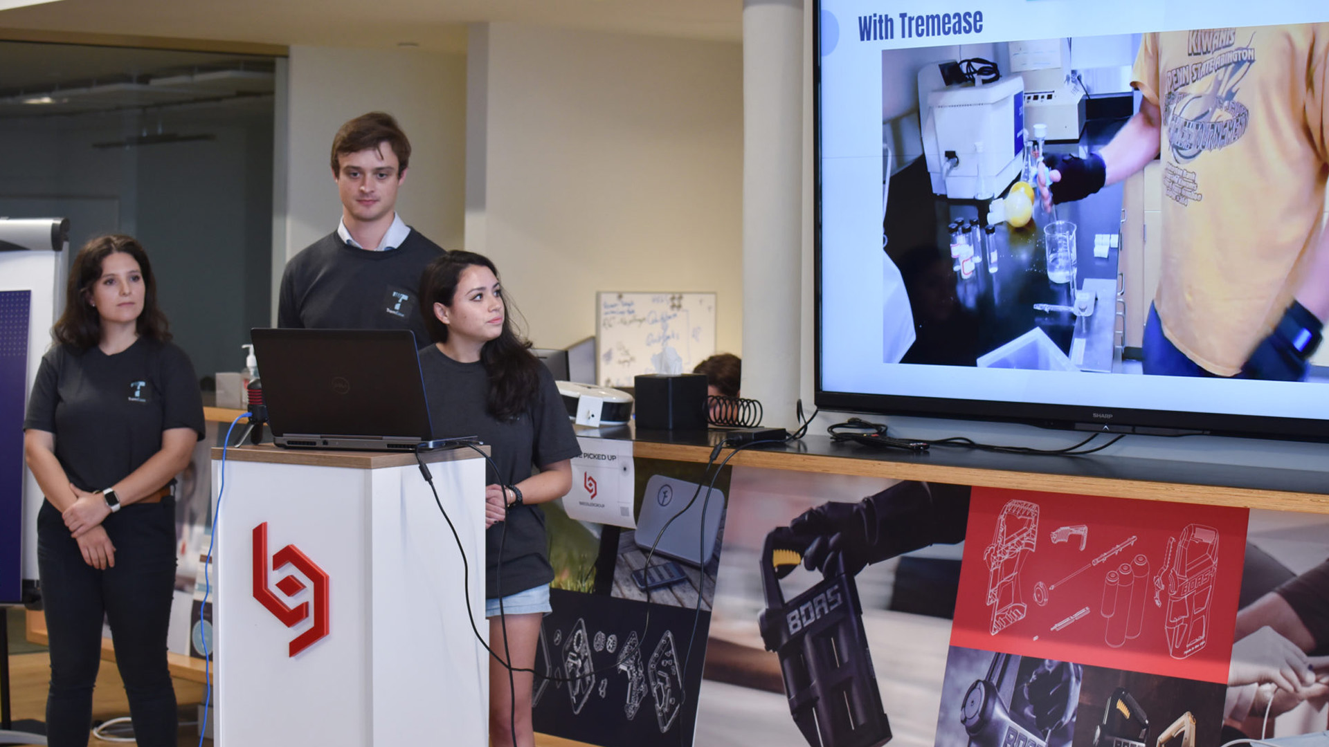 TremEase creators stand at a podium presenting with a laptop while a screen behind them displays their weighted glove design concept.