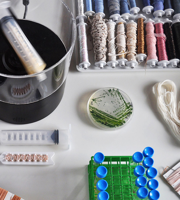 An overhead view of various dyes, colorful yarns/thread and black and white embroidered fabrics sitting on a white surface.