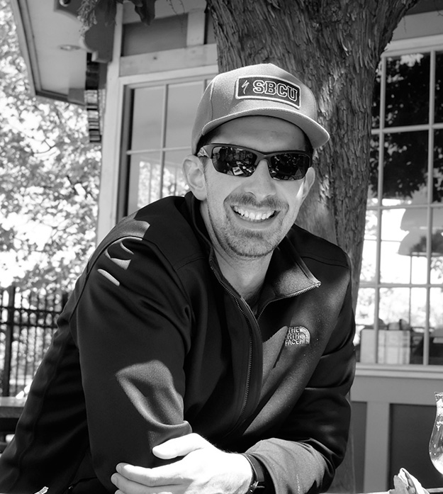 A black-and-white photo of Eli Tuttle smiling at the camera while wearing a hat and sunglasses as he leans forward on a table outside.