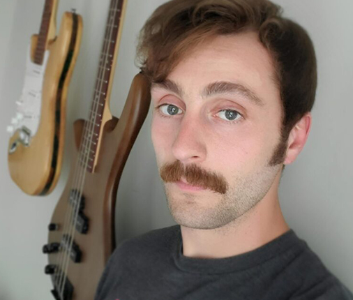 Isaac Savinese sports a mustache while wearing a T-shirt and looking at the camera with two guitars hanging on the wall over his shoulder.