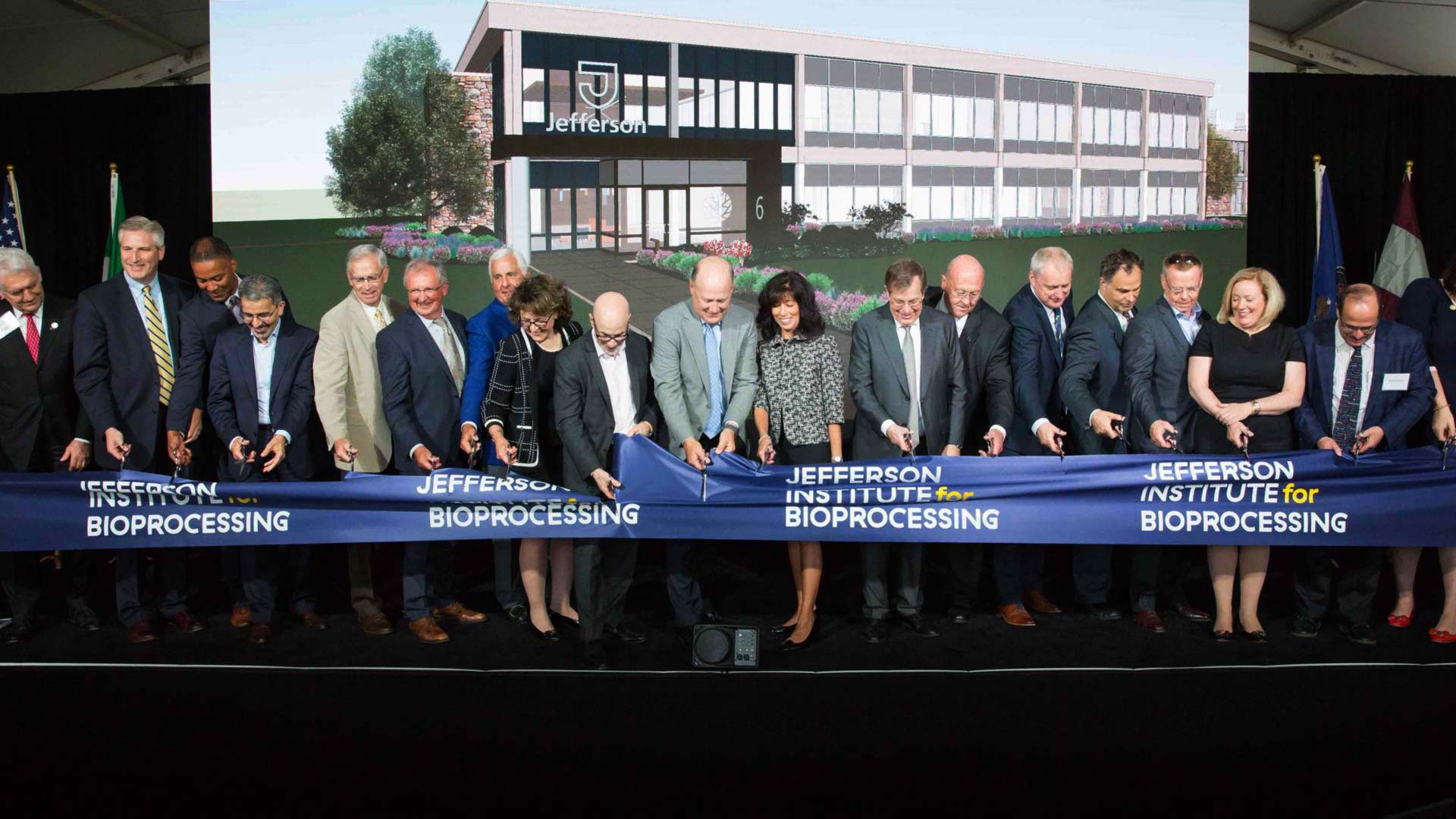 A group of people wearing formal business attire stand together with scissors in their hands as they cut a large blue ribbon in front of them that reads, “Jefferson Institute for Bioprocessing.”