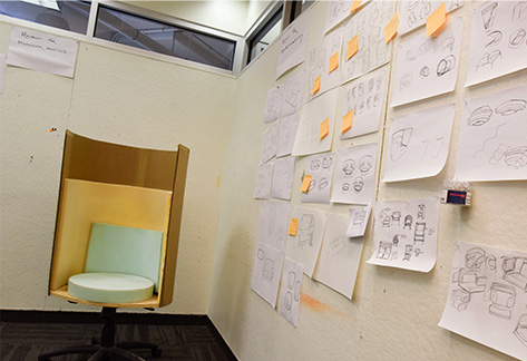 A corner of a room where one wall is covered in pieces of paper depicting chair concept sketches while the chair prototype sits in front of the other wall.