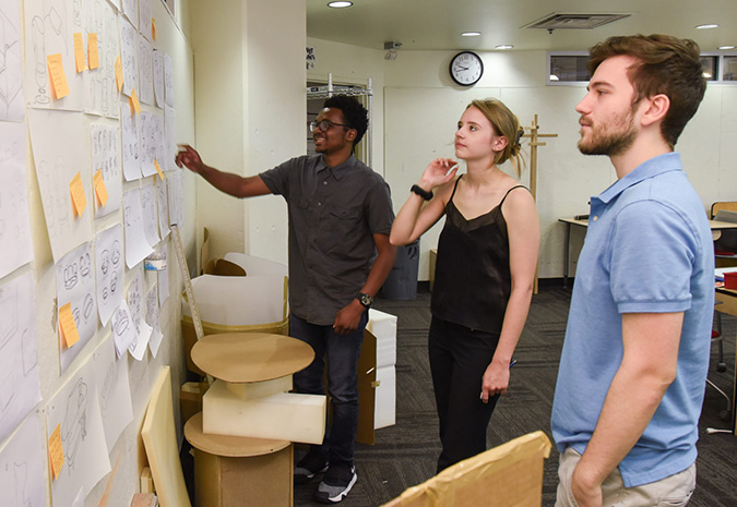 Three design team members stand side-by-side while looking at a wall covered in chair concept sketches.