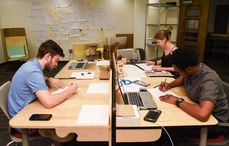 Three design team members sit at a large table covered in papers and laptops while drawing sketches together.