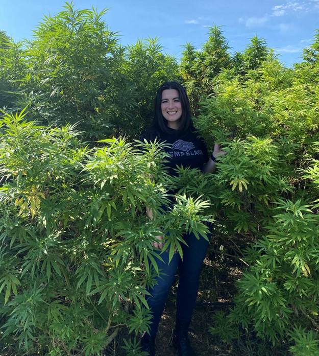 Isabella Siravo stands outside on a sunny day surrounded by hemp plants while smiling at the camera.