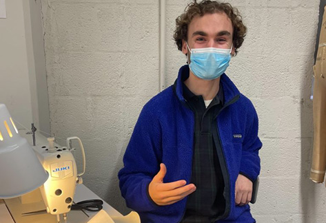 Jefferson student Arthur Hall sits behind a sewing machine while wearing a surgical mask, blue fleece jacket and khaki pants.