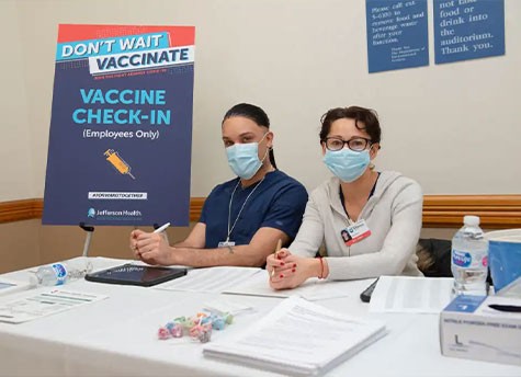 Employees Edward Davis and Betsy Rivera sitting at the table of the COVID-19 vaccine check-in at Thomas Jefferson University Hospital