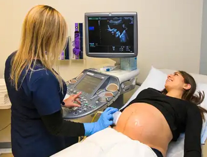 An expectant mother lies down in an exam room smiling at a screen while receiving a sonogram from a care provider.