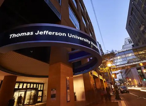 The exterior of a building in downtown Philadelphia that reads “Thomas Jefferson University Hospital” above the doorway and “Jefferson - Home of Sidney Kimmel Medical College” on the skyway spanning above the city street.